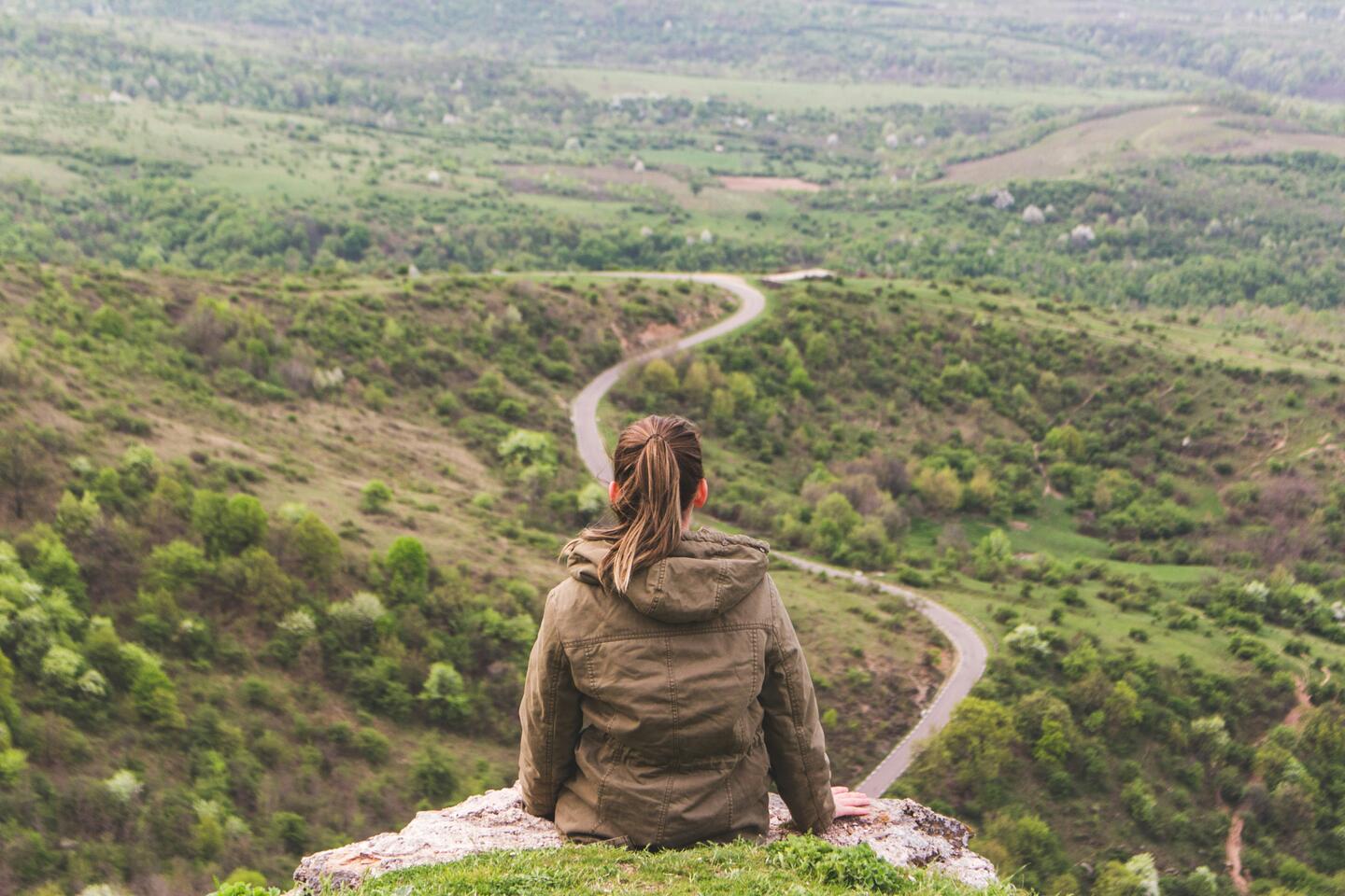 Femme assise sur une falaise, regard tourné vers une vallée traversée d'une route sinueuse - traverser un changement professionnel